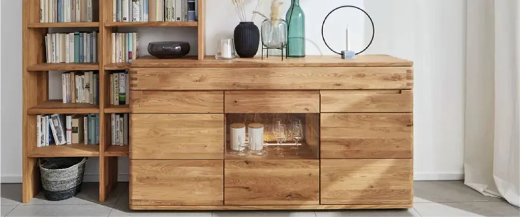 Hochwertiges Sideboard aus Massivholz mit integrierter Glasvitrine und dekorativen Vasen. Daneben ein Bücherregal aus Holz. Moderne Wohnmöbel in natürlicher Holzoptik für stilvolle und langlebige Wohnzimmer Einrichtung.
